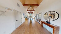 Located at 409 E William Cannon Dr in Austin, TX, this single-floor space offers a well-appointed, open layout that makes a strong first impression. The architecture is defined by dramatic vaulted ceilings with warm, exposed wood beams and tongue-and-groove paneling, lending the interior a sense of height and character throughout.

The main entrance hall is expansive and bright, anchored by a striking floor-to-ceiling pentagonal window above double glass doors that frame views of an outdoor pool area. Natural light pours in from multiple large windows, enhancing the airy, welcoming atmosphere. Light-toned hardwood flooring runs throughout, tying each area together cohesively.

The open central area transitions seamlessly into a workspace with a central island-style desk and bar-height seating, alongside a secondary gathering room complete with high-top stools and decorative wall art. A well-appointed lounge area features comfortable sofas, a patterned area rug, and access to a compact kitchenette with white upper cabinetry — ideal for hosting guests or residents.

Crisp white walls provide a clean backdrop for thoughtfully placed artwork and decorative accents, including woven wall pieces and sculptural elements. The overall ambiance is modern yet warm, balancing professional functionality with inviting residential comfort — making this a distinctive and well-designed community space.