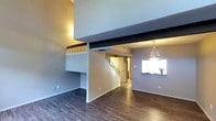 This two-story space showcases an open and airy layout defined by soaring ceilings that create a dramatic sense of vertical volume. A loft-style upper level overlooks the main living area below, with a wood-railed balcony adding architectural character and visual interest. Dark hardwood-style flooring runs throughout the main level, providing a rich contrast against the light gray walls. A prominent exposed beam spans the ceiling, reinforcing the industrial-meets-modern aesthetic of the space. The staircase, featuring classic wood balusters, connects the two levels and is centrally positioned, making it a natural focal point. A pass-through window opening toward what appears to be a kitchen area adds functional connectivity between spaces, while a modern geometric pendant light fixture above hints at a dedicated dining area. The overall ambiance is clean and contemporary, with neutral tones and thoughtful architectural details contributing to a spacious, well-structured interior that balances openness with defined functional zones across both levels.