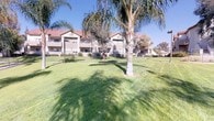 This residential community showcases a Spanish-inspired architectural style, characterized by tan stucco exteriors, distinctive red tile rooflines, and clean white-trimmed windows. The multi-story buildings are arranged to face generously sized, well-maintained common green spaces that serve as the visual centerpiece of the property. Expansive lawns stretch between the structures, lined with mature palm trees, shade trees, and ornamental plantings that contribute to a lush, park-like atmosphere.

Each building features private balconies with wrought iron railings on the upper levels, providing residents with outdoor space and views of the landscaped grounds. Ground-floor units have access to patio areas bordered by low fencing and native plantings. The exterior walls are finished in smooth stucco, and exterior lighting fixtures are mounted near windows and entryways for practical illumination.

Dedicated central HVAC units are positioned along the building exteriors, ensuring climate control for each unit. Perimeter fencing and gated access points are visible throughout the grounds, contributing to the community's sense of security. The overall setting, benefiting from abundant natural sunlight and a well-groomed landscape, creates a comfortable and inviting outdoor environment suited to warm-weather living.