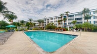This single-floor residence is part of a well-appointed multi-story building designed in a tropical coastal style. The exterior of the complex is finished in crisp white with blue shutters and trim, evoking a classic Florida aesthetic. Tall palm trees line the building's façade, reinforcing the warm, resort-inspired atmosphere.

A standout feature of the complex is the expansive outdoor pool, which serves as the centerpiece of the shared amenity space. The generously sized pool is surrounded by a broad paved deck furnished with lounge chairs and seating areas, providing ample space for relaxation. Lush tropical plantings and landscaping borders frame the pool area, adding color and privacy. Residents of the building have direct access to this outdoor retreat, which is clearly visible from the building's multiple balconies above.

While interior images were not sufficiently lit to provide detailed room-by-room descriptions, the unit is situated on a single level within the complex. The building's design suggests that units may feature private balconies with views overlooking the pool and surrounding greenery, contributing to an overall ambiance that is relaxed, sun-drenched, and reminiscent of a coastal retreat.