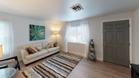 This single-floor space is thoughtfully laid out to maximize functionality and comfort across its main living areas. The living room serves as the heart of the unit, featuring light hardwood-style flooring, neutral walls, and natural light streaming through windows dressed with sheer white curtains and blinds. A striped area rug anchors the seating area, complemented by a cream sofa and decorative accents including a bold botanical canvas. A dedicated media area is outfitted with a dark wood console and wall-mounted artwork.

A separate sitting nook adjoins the living space, featuring a decorative round mirror, glass side table, and accent chair, offering an inviting secondary lounge area.

The kitchen is well-equipped with warm wood cabinetry, ample counter space, and black appliances including a range, dishwasher, and refrigerator. Light flooring ties it visually to the rest of the home.

The bedroom is carpeted for warmth, with windows allowing soft natural light. It comfortably accommodates a queen-sized bed flanked by dark wood nightstands.

The bathroom includes a tub-shower combination with a curtain, a white vanity with sink, and clean tile finishes throughout, providing a functional and tidy space.