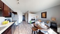 This single-floor space showcases a thoughtfully designed open-concept layout that seamlessly integrates the kitchen, dining, and living areas into one cohesive living environment. The kitchen is outfitted with sleek dark cabinetry, stainless steel appliances — including a refrigerator, stove, and built-in microwave — and provides efficient use of space without compromising functionality. A round wooden dining table with industrial-style bar stools anchors the dining area, creating an intimate setting ideal for everyday meals or entertaining. The living area flows naturally from the dining space, offering a comfortable seating arrangement accented by decorative artwork, including a prominent framed reproduction of Gustav Klimt's "The Kiss," and warm ambient lighting provided by a contemporary floor lamp. Throughout the main living space, smooth hardwood-style flooring adds warmth and visual continuity. The neutral gray-toned walls and white ceilings contribute to a clean, modern aesthetic, while recessed and track lighting ensure the space is well-lit. A hallway toward the rear leads to additional private areas, including what appears to be a bedroom and bathroom. The overall ambiance is modern, functional, and inviting.