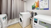 Located at 1050 Highland St in Burlington, Ontario, this shared laundry facility offers a clean, functional, and well-organized space for residents. The room is finished with durable light ceramic tile flooring and bright white walls, creating a tidy and practical environment.

The main area houses a set of coin-operated washers and dryers, including both front-loading and top-loading machines, all clearly labeled with posted usage guidelines and laundry detergent instructions for user convenience. A continuous grey laminate folding counter runs along one wall, providing ample surface space for sorting and folding laundry. Exposed ventilation ductwork services the dryers efficiently along the back wall.

A coin payment kiosk is conveniently positioned near the entrance, streamlining the payment process. The space also includes a closet, ideal for storing laundry-related supplies. Colorful abstract artwork displayed on the walls adds a welcoming touch to the utilitarian setting. A curtained window provides a degree of privacy while allowing natural light to filter in.

Overall, this laundry room is a well-maintained, thoughtfully arranged shared facility that meets the everyday laundry needs of building residents with ease and practicality.