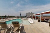 Located at 600 St Clair Ave SW in Huntsville, Alabama, this rooftop pool deck offers an impressive outdoor retreat with commanding views of the city and the rolling mountains beyond. The centerpiece of the space is a generously sized swimming pool finished with aqua-toned tile, surrounded by a broad stone-paved deck that provides ample room for relaxation and recreation. Dozens of modern lounge chairs and chaise lounges are arranged throughout the deck, offering guests a variety of spots to sunbathe or unwind. A stylish pergola structure with warm wood accents and retractable red canopy panels anchors one side of the deck, providing shaded seating areas with a more intimate ambiance. Raised planter beds with lush greenery add a touch of nature to the urban setting, while umbrella-shaded dining tables offer additional outdoor seating options. The open-air design ensures abundant natural light throughout the day, and the elevated position affords unobstructed panoramic views, including the iconic church steeple of the Huntsville skyline. The clean, contemporary aesthetic of the deck, combined with its thoughtful layout and varied amenities, makes this rooftop pool area a standout feature of the property.