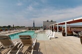 Located at 600 St Clair Ave SW in Huntsville, Alabama, this rooftop pool deck offers an impressive outdoor retreat with commanding views of the city and the rolling mountains beyond. The centerpiece of the space is a generously sized swimming pool finished with aqua-toned tile, surrounded by a broad stone-paved deck that provides ample room for relaxation and recreation. Dozens of modern lounge chairs and chaise lounges are arranged throughout the deck, offering guests a variety of spots to sunbathe or unwind. A stylish pergola structure with warm wood accents and retractable red canopy panels anchors one side of the deck, providing shaded seating areas with a more intimate ambiance. Raised planter beds with lush greenery add a touch of nature to the urban setting, while umbrella-shaded dining tables offer additional outdoor seating options. The open-air design ensures abundant natural light throughout the day, and the elevated position affords unobstructed panoramic views, including the iconic church steeple of the Huntsville skyline. The clean, contemporary aesthetic of the deck, combined with its thoughtful layout and varied amenities, makes this rooftop pool area a standout feature of the property.