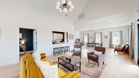 This single-floor space is thoughtfully designed with an open, airy layout that seamlessly connects its primary functional zones. The central living area is anchored by soaring vaulted ceilings and a striking multi-globe chandelier, creating an immediate sense of grandeur. Light hardwood flooring runs throughout, contributing to the cohesive, modern aesthetic. The lounge area features a bold dark teal accent wall, providing visual contrast against the otherwise crisp white walls, and is furnished with warm mustard-toned seating and a mid-century-inspired bookshelf.

Adjacent to the lounge, a pass-through kitchen bar with counter seating connects the social areas to a galley-style kitchen. The kitchen is outfitted with white shaker cabinetry, gold-toned hardware, quartz countertops, a classic white subway tile backsplash, and stainless steel appliances—all set against dramatic dark accent walls.

The dining and gathering area occupies a generously proportioned space filled with natural light from multiple windows dressed with linen curtains. A large wooden dining table with seating for several guests serves as the focal point, complemented by a television console along one wall. A private office nook features deep slate-blue walls and a dedicated workspace. Direct exterior access is available through glass doors, connecting the interior to an outdoor area.