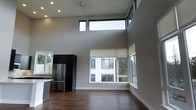 This single-floor space showcases a striking contemporary design defined by dramatically high ceilings that create an airy, open atmosphere. The living area benefits from an abundance of natural light, drawn in through a combination of large picture windows at eye level and a row of clerestory windows positioned near the ceiling, ensuring consistent brightness throughout the day. The black-framed windows provide a bold contrast against the neutral gray walls, adding a modern architectural edge to the interior.

The open-concept layout connects the living space directly to the kitchen, which features dark upper cabinetry, a stainless steel refrigerator, and light-toned countertops. This contrast of dark and light finishes contributes to a sleek, contemporary aesthetic. Rich, dark hardwood flooring runs throughout, grounding the space and adding warmth against the cool gray palette.

A ceiling fan with recessed lighting provides both ventilation and illumination, complementing the natural light. The overall design emphasizes clean lines, generous vertical space, and a minimalist sensibility, making this a well-appointed and visually compelling modern living environment.