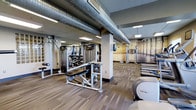This fitness center offers a thoughtfully designed workout environment that caters to a wide range of exercise needs. The space features an exposed industrial ceiling with large ventilation ductwork, lending a modern and utilitarian aesthetic that is complemented by recessed and panel lighting throughout. The flooring consists of durable, multi-toned carpet tiles well-suited for a high-traffic fitness environment.

The strength training area is generously outfitted with a variety of cable machines, weight benches, and multi-functional resistance equipment, providing options for both isolated and compound movements. A dedicated dumbbell rack adds to the free weights selection available. Precor-branded cardio equipment is also present, reflecting a commitment to quality and performance.

Structural columns are integrated throughout the space and are finished to match the overall color palette of grays and neutrals, contributing to a cohesive design. A large-scale photographic mural adorns one wall, depicting a fitness-themed scene that adds visual depth and motivation to the space. The layout is organized to allow for comfortable circulation between equipment stations, supporting a practical and efficient workout experience from start to finish.