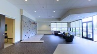 This single-floor multi-purpose space is thoughtfully designed to balance active recreation with relaxed social interaction. The most striking feature is a large climbing wall adorned with colorful handholds, positioned alongside padded activity mats that provide a safe and functional workout zone. A basketball hoop mounted at height adds to the athletic character of the space.

The lounge area offers a welcoming contrast to the active zone, furnished with a spacious sectional sofa and accent chairs arranged to encourage conversation and relaxation. Large windows with roller shades line the perimeter, flooding the interior with natural light and offering views of the surrounding outdoor environment.

The interior finishes are clean and modern, with a drop ceiling featuring recessed lighting that ensures consistent illumination throughout. The durable rubberized flooring is well-suited to both physical activity and everyday foot traffic. The open-plan layout allows the space to serve a variety of uses simultaneously, making it an ideal environment for community engagement, recreational programming, or informal gatherings. Overall, the space strikes a practical yet inviting tone, designed to accommodate diverse groups and activities with ease.