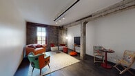 This single-floor loft-style space masterfully combines industrial heritage with modern design sensibilities. The living area serves as the centerpiece, showcasing original exposed brick walls that span from floor to ceiling, adding warmth and historic character. Substantial reclaimed timber beams and a structural wooden post anchor the space overhead, while track lighting illuminates the room from above. Dark hardwood flooring runs throughout, grounding the eclectic, colorful furnishings arranged on a patterned area rug. A tall window allows natural light to filter in, framing outdoor views.

The bedroom continues the industrial aesthetic, retaining exposed timber beams on the ceiling and original brick detailing. A lean black metal shelving unit and a full-length oval mirror complement the space, which benefits from track lighting overhead.

The kitchen presents a clean contrast with its modern, minimalist design. White upper and lower cabinetry pairs with white quartz countertops, a subway-tiled backsplash, and a full suite of stainless steel appliances, including a range, dishwasher, and refrigerator.

The bathroom maintains a fresh, contemporary feel with large-format tile flooring, a white floating vanity with ample storage, and a tub-shower combination. Overall, the space achieves a compelling balance between raw architectural character and polished modern functionality.