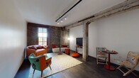 This single-floor loft-style space masterfully combines industrial heritage with modern design sensibilities. The living area serves as the centerpiece, showcasing original exposed brick walls that span from floor to ceiling, adding warmth and historic character. Substantial reclaimed timber beams and a structural wooden post anchor the space overhead, while track lighting illuminates the room from above. Dark hardwood flooring runs throughout, grounding the eclectic, colorful furnishings arranged on a patterned area rug. A tall window allows natural light to filter in, framing outdoor views.

The bedroom continues the industrial aesthetic, retaining exposed timber beams on the ceiling and original brick detailing. A lean black metal shelving unit and a full-length oval mirror complement the space, which benefits from track lighting overhead.

The kitchen presents a clean contrast with its modern, minimalist design. White upper and lower cabinetry pairs with white quartz countertops, a subway-tiled backsplash, and a full suite of stainless steel appliances, including a range, dishwasher, and refrigerator.

The bathroom maintains a fresh, contemporary feel with large-format tile flooring, a white floating vanity with ample storage, and a tub-shower combination. Overall, the space achieves a compelling balance between raw architectural character and polished modern functionality.