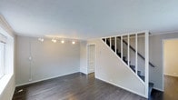 This three-floor space offers a straightforward and functional layout with a clean, neutral design aesthetic. The main living area is highlighted by dark hardwood flooring that contrasts crisply against bright white baseboards, trim, and walls painted in a soft, light grey tone. A track lighting fixture runs along the ceiling, providing adjustable, directed illumination across the open room.

Crown molding lines the ceiling, adding a subtle architectural detail that elevates the overall finish of the space. A white paneled interior door provides access to an additional room or utility area, contributing to the practical layout of this level.

A white-railed staircase with dark treads is prominently positioned within the main living area, connecting this floor to the levels above and below. The staircase features a clean open-rail design with vertical white balusters, maintaining the consistent light color palette found throughout the space.

The overall ambiance is minimal and move-in ready, with neutral tones and simple finishes that offer flexibility for a variety of interior styles. The three-floor configuration provides ample vertical separation of living spaces, making it a well-structured and versatile environment.