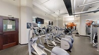 This fitness center at 801 Pine St in St. Louis, Missouri, spans a generously sized single floor, providing ample room for a comprehensive workout experience. The space is divided into two distinct functional zones: a cardio area and a strength training area, together offering a full range of exercise options.

The cardio zone features a lineup of Precor elliptical trainers, treadmills, and stationary bikes, all positioned along a wall of floor-to-ceiling mirrors with mounted flat-screen televisions. The mirrors enhance the sense of space while allowing users to monitor their form. Overhead, industrial-style linear lighting illuminates the area brightly, complementing the natural light that filters in through windows along the perimeter.

The strength training area is anchored by a bold orange accent wall that adds a vibrant, energetic atmosphere to the space. This section is equipped with a variety of cable machines, weight benches, and functional training equipment. Exposed ductwork and high ceilings reinforce the industrial character of the facility, while durable rubber flooring throughout ensures safety and comfort.

Additional amenities include a water station, shelving for accessories, and wall-mounted TVs throughout. The layout is open and well-organized, allowing for efficient traffic flow between equipment. The overall design balances industrial aesthetics with a clean, functional environment suited for a full-body fitness regimen.