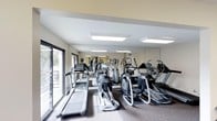 This fitness center is designed to provide a functional and motivating workout environment. The space is outfitted with a comprehensive selection of cardio equipment, including treadmills, elliptical trainers, and stair climbers, all arranged to maximize floor space and ease of movement. A full-length mirrored wall runs along one side of the room, visually expanding the space and allowing users to monitor their form during exercise. The flooring features a durable, low-pile carpet well-suited for a high-traffic fitness environment. Overhead, recessed panel lighting provides bright, even illumination throughout the room. The angled ceiling adds an architectural dimension to the space, contributing to an open and airy feel. Large sliding glass doors along one wall flood the interior with natural light and offer views of the surrounding greenery, creating a refreshing connection to the outdoors. The clean, neutral color palette of white and light gray walls contributes to a crisp and professional atmosphere. Overall, the fitness center is thoughtfully laid out and well-maintained, offering an inviting and practical setting for daily exercise routines.