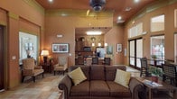This inviting single-floor community space is designed with a cohesive Southwestern-inspired aesthetic, characterized by rich terracotta and warm adobe tones throughout. The main living area is generously proportioned, featuring soaring ceilings, a ceiling fan, and large windows dressed with wood blinds that filter natural light across the stone tile flooring. Comfortable lounge seating, framed artwork, and lush potted plants — including a striking full-height ficus tree — lend a welcoming, residential feel to the space.

The open-concept kitchen is fully equipped with stainless steel appliances, including a side-by-side refrigerator, a gas range, and a built-in microwave. Rich dark wood cabinetry extends to the ceiling, accented by decorative wicker baskets and greenery. A spacious countertop island serves as a natural gathering point between the kitchen and living area.

A standout feature is the dramatic rotunda dining room, enclosed by a curved wall of floor-to-ceiling windows that flood the space with natural light and provide views of the surrounding outdoor areas. High clerestory windows further elevate the room's sense of openness. Dark wood bistro tables and upholstered chairs create an intimate yet airy dining environment, making this a functional and stylish gathering space overall.