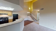 Located at 12700 Bloomfield Ave in Norwalk, California, this two-story residence offers a well-organized layout with thoughtful design details throughout. The main level opens into a spacious living area boasting dramatically high ceilings and a striking double-height wall that creates an airy, open atmosphere. Warm-toned carpet flows through the living space, while durable wood-look flooring defines the kitchen and entry areas. A staircase with white railings and a pendant chandelier serves as a stylish focal point connecting both levels. The kitchen is well-appointed with light wood cabinetry, granite-style countertops, a full suite of black appliances including a gas range and refrigerator, and a skylight that floods the space with natural light. A peninsula counter provides additional workspace and casual seating potential. The upper level features bedrooms with vaulted ceilings, generous closet space with mirrored sliding doors, and windows fitted with blinds that invite natural light. One bedroom opens to a private balcony with outdoor views of the surrounding neighborhood. The bathrooms are clean and functional, featuring tub-shower combinations, vanities with white countertops, and wood-look flooring. Overall, the residence offers a comfortable and practical living environment with notable architectural volume and well-distributed natural light.