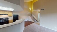 Located at 12700 Bloomfield Ave in Norwalk, California, this two-story residence offers a well-organized layout with thoughtful design details throughout. The main level opens into a spacious living area boasting dramatically high ceilings and a striking double-height wall that creates an airy, open atmosphere. Warm-toned carpet flows through the living space, while durable wood-look flooring defines the kitchen and entry areas. A staircase with white railings and a pendant chandelier serves as a stylish focal point connecting both levels. The kitchen is well-appointed with light wood cabinetry, granite-style countertops, a full suite of black appliances including a gas range and refrigerator, and a skylight that floods the space with natural light. A peninsula counter provides additional workspace and casual seating potential. The upper level features bedrooms with vaulted ceilings, generous closet space with mirrored sliding doors, and windows fitted with blinds that invite natural light. One bedroom opens to a private balcony with outdoor views of the surrounding neighborhood. The bathrooms are clean and functional, featuring tub-shower combinations, vanities with white countertops, and wood-look flooring. Overall, the residence offers a comfortable and practical living environment with notable architectural volume and well-distributed natural light.