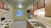 This single-floor residential space offers a practical and functional layout designed to maximize everyday comfort. The kitchen is a central feature, equipped with warm wood-toned cabinetry spanning both upper and lower sections, providing generous storage throughout. A double-basin stainless steel sink sits beneath overhead cabinets, while a countertop range and range hood add to the kitchen's full functionality. The light-toned countertops offer ample workspace, making the area well-suited for meal preparation. The kitchen flows openly into an adjoining living area, which features neutral carpet flooring that adds warmth to the space. A window dressed with vertical blinds allows natural light to filter in, brightening the room and offering a view of surrounding greenery outdoors. A baseboard heating unit runs along the wall beneath the window, ensuring comfortable temperatures throughout the year. The overall color palette is soft and neutral, with light green walls creating a calm and understated ambiance. Flush ceiling lighting provides consistent illumination across both the kitchen and living areas. The open-concept connection between the kitchen and living space makes the layout feel cohesive and easy to navigate, offering a straightforward and comfortable living environment.