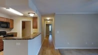 This single-floor residence offers a thoughtfully designed layout with a clean, functional aesthetic throughout. The interior is anchored by warm hardwood flooring that extends through the main living and hallway areas, transitioning to carpet in the bedroom zones. A standout feature is the kitchen, which includes rich wood cabinetry and a granite-topped breakfast bar that opens toward the main living space, creating an inviting, open feel between the kitchen and the rest of the home. Flush-mounted ceiling fixtures provide consistent, warm lighting throughout. Crown molding adds a refined architectural detail to the main living area, contributing to an overall polished look. The central hallway runs the length of the home, connecting the main living spaces to the private rooms, with natural light visible at the far end near the entryway. Neutral, warm-toned walls throughout create a versatile backdrop that complements a variety of interior styles. The space is practical in its design, with visible utility features such as ceiling vents and light switches discreetly integrated. Overall, the residence presents a comfortable, move-in-ready environment with classic finishes and a straightforward, accessible floor plan.