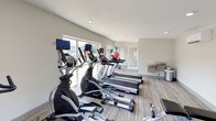 This fitness center offers a clean, modern environment designed to support a well-rounded workout routine. The space is finished with light wood-look flooring and neutral gray walls, creating a bright and energizing atmosphere throughout. Recessed lighting complements the abundant natural light that streams in through large windows and glass doors lining multiple walls, providing open views of the surrounding outdoor areas, including a pool and landscaped grounds.

The cardio section is well-stocked with a row of Spirit-brand treadmills and elliptical cross-trainers, each equipped with individual monitors for entertainment during workouts. A dedicated dumbbell rack along one wall offers a range of free weights for strength training, accompanied by a weight bench for added versatility. Exercise and medicine balls in various sizes are also available, supporting flexibility and functional fitness routines. Additional strength and conditioning equipment rounds out the offerings.

A utility counter along one wall provides a convenient space for water or personal belongings during workouts. The open layout ensures comfortable circulation between equipment, and the glass door access offers easy entry and exit. The overall ambiance is light, airy, and purposefully functional, making this fitness center a practical and inviting space for daily exercise.