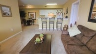 This single-floor space showcases an open-concept layout that seamlessly connects the living area, dining room, and kitchen, creating a cohesive and functional environment. The living area features plush wall-to-wall carpeting and is anchored by a dark wood coffee table paired with a comfortable brown upholstered sofa, offering a welcoming atmosphere for relaxation. Warm yellow-toned walls add a sense of warmth throughout the space.

The kitchen is positioned at the center of the layout, highlighted by a raised breakfast bar with elegant wrought-iron barstools that provide casual seating for three. White cabinetry lines the kitchen walls, complemented by standard appliances including a refrigerator and microwave. The bar counter serves as a natural divider between the kitchen and the living area.

Adjacent to the kitchen, the dining area accommodates a dark wood table surrounded by matching chairs, ideal for seated meals. Track lighting and overhead fluorescent fixtures ensure the space is well-lit. A decorative large plant and framed wall art add character, while the open floor plan contributes to an airy and spacious overall ambiance.