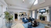 This single-story space makes a strong first impression with its dramatic vaulted ceilings that draw the eye upward, accentuated by recessed lighting and a transom window that introduces natural light from above. The main living area is thoughtfully arranged with plush navy blue armchairs, a patterned area rug in coordinating blues, and a dark wood coffee table, creating an inviting and cohesive aesthetic. A stone-surround fireplace anchors one wall, adding warmth and a classic architectural focal point to the room. Bold patterned drapery flanks the windows, complementing the blue and white color scheme throughout. An elegant arched passageway connects the lounge to a kitchen bar area, which features light cabinetry, bar-height seating, and appliances including a full-size refrigerator — suggesting the space supports events or gatherings requiring refreshments. The flooring transitions between warm travertine tile and area rugs, while wainscoting and clean white walls lend a polished, finished quality. A wall-mounted flat-screen television adds a modern, functional touch to the overall design.