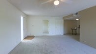 This single-floor space presents an open and functional layout finished in neutral tones throughout. The main living area is generously proportioned, featuring wall-to-wall carpet in a light gray hue that extends across the entire floor, contributing to a clean and understated aesthetic. A ceiling fan with an integrated light fixture is centrally mounted, providing both air circulation and ambient lighting to the space.

The walls are painted in a soft, warm white, complemented by a textured ceiling that adds subtle visual interest. Natural light enters through a large window positioned along the left-hand wall, brightening the interior without the need for additional lighting during the day. A small entry mat near the window suggests a secondary access point.

Toward the rear of the main room, an open doorway leads into an adjacent room, offering additional usable space. A second door, likely a closet, is positioned nearby. To the right, a partially separated area — defined by a subtle change in wall color — provides a distinct zone that could serve as a dining or workspace, and appears to accommodate a small piece of furniture. Ventilation is supported by a visible ceiling vent in this section, indicating a functional HVAC system throughout.