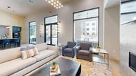 Located at 185 Marquis Way in Freeport, Florida, this thoughtfully designed single-floor amenity space offers a well-appointed environment ideal for both socializing and productivity. The open-concept kitchen and living area serves as the heart of the space, anchored by a striking quartz island with waterfall edges, navy blue cabinetry, and a shimmering penny-tile backsplash accented by a bold branded medallion. A full-size stainless steel refrigerator and sleek pendant lighting complete the kitchen's polished aesthetic. The adjacent living area boasts soaring ceilings, floor-to-ceiling windows that flood the room with natural light, and views of the surrounding outdoor amenities. A statement fireplace clad in textured tile with a wall-mounted TV serves as the focal point of the lounge seating area, complemented by contemporary furnishings and a designer area rug. A cluster pendant chandelier adds warmth and visual interest overhead. The dedicated office and conference rooms feature sage green accent walls, large glass partitions, wall-mounted displays, built-in navy cabinetry, and vibrant geometric rugs, creating a professional yet inviting atmosphere. Light wood-look flooring flows throughout, tying together the cohesive and modern design of this versatile community space.