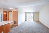 This single-floor space offers a functional and open layout with several notable design elements. The kitchen serves as a central hub, featuring a wraparound peninsula with a light countertop, warm wood-toned lower cabinetry, white upper cabinets, and a full suite of appliances including a refrigerator, electric range, and microwave. An elegant chandelier overhead connects the kitchen seamlessly to the adjacent dining area, enhancing the open, connected feel of the shared space.

The living area is generously sized, finished with plush carpeting, and benefits from vaulted ceilings that add a sense of openness and volume. A sliding glass door with vertical blinds provides direct access to an outdoor area and allows for abundant natural light. Additional interior doors lead to closets or utility spaces near the main entry.

A separate carpeted room with a large window fitted with horizontal blinds offers a bright and versatile space, ideal as a bedroom or home office. The vaulted ceiling continues here, reinforcing the airy character of the unit.

The bathroom is well-appointed, featuring a full tub-shower combination, a large vanity with white cabinetry and countertop, an expansive mirror with overhead lighting, and durable wood-look flooring that adds a clean, modern contrast to the neutral walls.