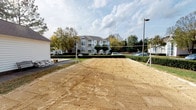 This outdoor recreational amenity space offers two distinct sport courts designed for active community use. The sand volleyball court is equipped with a regulation net and posts, set on a leveled sand surface with defined boundaries. The surrounding landscape features trimmed hedges and mature trees that provide a natural backdrop, while dedicated lighting poles ensure the court remains usable into the evening hours.

Adjacent to the volleyball area is a full-size tennis court finished in a classic green hard-court surface with crisp white boundary lines and a center net. The court is fully enclosed by a green chain-link fence with a dedicated entry gate, offering a secure and focused playing environment. Overhead court lighting is installed to support extended play beyond daylight hours.

The grounds surrounding both courts are thoughtfully landscaped with established shrubs, decorative mulch beds, and a variety of trees showcasing seasonal foliage. A multi-story residential building is visible in the background, contextualizing these courts as shared community amenities. The overall layout is open and well-maintained, creating an inviting and functional outdoor sports environment suitable for residents of all skill levels.