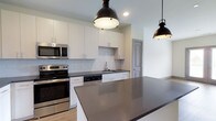 This single-floor space showcases a thoughtfully designed, modern kitchen that serves as its centerpiece. The kitchen features crisp white shaker-style cabinetry that extends to the ceiling, providing generous storage throughout. Dark charcoal countertops offer a striking contrast against the white cabinetry, while a classic subway tile backsplash adds texture and a clean, timeless aesthetic. The kitchen is equipped with a full suite of stainless steel appliances, including a double wall oven, built-in microwave, and a dishwasher, making it both functional and stylish. A large central island with matching dark countertop provides ample workspace and serves as a natural gathering point. Overhead, a pair of matte black industrial-style dome pendant lights add a bold, contemporary accent while providing focused task lighting. Recessed lighting supplements the pendants for well-distributed illumination throughout the space. Light wood-tone flooring grounds the room warmly, complementing the cool tones of the cabinetry and countertops. The open layout allows for a seamless flow between the kitchen and the adjoining living areas, creating an inviting and highly functional environment suited for both everyday living and entertaining.