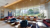 This single-floor space presents a thoughtfully designed commercial interior that balances refined aesthetics with practical functionality. The reception area greets visitors with large-format light stone tile flooring, a sculptural wood reception desk, a decorative sunburst wall fixture, and floor-to-ceiling glass walls that draw in abundant natural light and views of the surrounding landscaped grounds.

The main lounge area is defined by a dramatic wall of floor-to-ceiling glazing that frames views of mature flowering trees and manicured gardens, flooding the interior with daylight. Warm timber slatted ceilings run throughout, complementing a richly patterned mosaic carpet that anchors an eclectic mix of seating — including bold teal and red upholstered chairs, a built-in cushioned banquette, and contemporary side tables. A more intimate seating alcove features textured stone-clad walls, curated artwork, and layered soft furnishings.

The kitchenette and bar area offers a long dark-topped communal counter with bar seating, integrated appliances, open display shelving, and a textured copper-toned backsplash. Transition corridors feature travertine-clad columns and direct access to an outdoor courtyard, reinforcing the space's strong connection to the exterior environment throughout.