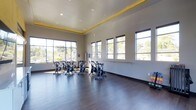 This single-floor fitness studio is designed to support a variety of exercise activities in a well-lit, open environment. The space is anchored by a row of spin bikes arranged across the dark hardwood flooring, providing ample room for cycling classes. Along one wall, a mounted ballet barre offers versatility for stretching, dance, or barre-based workouts.

The studio benefits from generous natural light, courtesy of large floor-to-ceiling windows along two walls that frame views of surrounding greenery. This abundance of natural light creates an energizing and inviting atmosphere. The neutral-toned walls complement the bright, airy feel of the space.

The ceiling features a distinctive tray design accented with yellow trim lighting, adding a modern, stylish touch to the room's overall aesthetic. Recessed lighting supplements natural light for evening or low-light sessions. The dark hardwood flooring provides both durability and a clean, polished appearance suited to high-traffic fitness use. The open layout ensures freedom of movement, making the studio functional for individual workouts or group fitness classes alike.