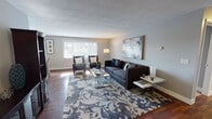 Located at 215 Greystone Ln in Rochester, NY, this single-floor residence offers a thoughtfully arranged layout with distinct living, sleeping, and functional spaces. The living room serves as the centerpiece of the home, featuring rich hardwood flooring, neutral gray walls, and a large picture window that draws in generous natural light and outdoor views. A boldly patterned area rug anchors the seating arrangement, which includes a substantial dark sofa, paired accent chairs, and a glass-topped coffee table, creating an inviting and contemporary atmosphere. A large abstract artwork adorns the wall, adding an artistic focal point to the room.

The kitchen is efficiently designed with warm-toned wood cabinetry, granite-style countertops, hardwood flooring, and white appliances, providing ample storage and workspace. The bedrooms are generously proportioned, featuring plush carpeting, neutral walls, and windows that admit natural light. The primary bedroom offers notable space, accommodating a large bed, mirrored nightstands, and additional seating. The bathrooms are finished with tile walls, mosaic tile flooring, rich wood vanities, and overhead lighting, delivering a clean and functional aesthetic throughout.