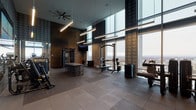 This upscale fitness center is designed to deliver a premium workout experience within a striking architectural setting. The space is defined by its soaring double-height ceilings, from which sleek linear pendant lights and a ceiling fan are suspended, creating an open, airy atmosphere. Floor-to-ceiling curtain-wall windows run along one side of the facility, flooding the interior with natural light and offering expansive panoramic views of the surrounding cityscape and beyond—a standout feature that distinguishes this gym from conventional fitness spaces.

The flooring throughout is finished in large-format rubber sport tiles in a neutral gray tone, providing both durability and comfort underfoot. Walls are dressed in a distinctive houndstooth-patterned tile treatment in varying scales, lending a refined, design-forward aesthetic. Mounted flat-screen televisions are positioned throughout for entertainment during workouts.

The facility is organized into distinct functional zones. The cardio area accommodates upright bikes, elliptical trainers, and a Peloton cycle. The strength and free weights section offers a full dumbbell rack backed by a mirrored wall, adjustable benches, a power rack with barbell and weight plates, cable machines, and a variety of selectorized equipment. A water station with dual drinking fountains and a towel disposal unit serves the space, and posted fitness rules ensure an orderly environment.