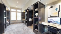 This single-floor space exudes a polished, professional ambiance through a thoughtful blend of rich materials and purposeful design. The interior is anchored by striking dark-stained built-in shelving units that rise from floor to ceiling, offering generous storage and display space for books, décor, and office essentials. The shelving serves as a natural room divider, creating distinct functional zones within the open layout while maintaining a sense of visual continuity throughout the space. Marble-style tile flooring in soft grey and taupe tones adds an air of sophistication underfoot, complementing the deep tones of the cabinetry. A built-in desk with a stone countertop provides a dedicated workspace, seamlessly integrated into the cabinetry design for a streamlined look. Large windows fitted with white blinds allow natural light to fill the interior, brightening the neutral wall tones and creating a welcoming atmosphere. Recessed lighting throughout ensures the space remains well-lit at any hour. The overall design balances elegance with practicality, making this a versatile environment well-suited for professional use, focused work, or a refined study setting.