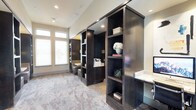 This single-floor space exudes a polished, professional ambiance through a thoughtful blend of rich materials and purposeful design. The interior is anchored by striking dark-stained built-in shelving units that rise from floor to ceiling, offering generous storage and display space for books, décor, and office essentials. The shelving serves as a natural room divider, creating distinct functional zones within the open layout while maintaining a sense of visual continuity throughout the space. Marble-style tile flooring in soft grey and taupe tones adds an air of sophistication underfoot, complementing the deep tones of the cabinetry. A built-in desk with a stone countertop provides a dedicated workspace, seamlessly integrated into the cabinetry design for a streamlined look. Large windows fitted with white blinds allow natural light to fill the interior, brightening the neutral wall tones and creating a welcoming atmosphere. Recessed lighting throughout ensures the space remains well-lit at any hour. The overall design balances elegance with practicality, making this a versatile environment well-suited for professional use, focused work, or a refined study setting.
