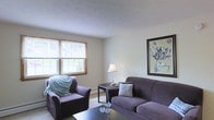 This single-floor residence offers a thoughtfully arranged living space designed for everyday comfort and functionality. The living room serves as the heart of the home, featuring neutral wall tones that create a calm, understated backdrop throughout. Plush, deep-toned upholstered seating — including a sofa and matching armchair — anchors the room, providing ample space to relax and unwind. A textured ceiling adds subtle architectural character overhead, while warm wood-trimmed windows along one wall invite natural light to filter in, offering pleasant views of the surrounding greenery outside. A classic table lamp contributes warm ambient lighting, complementing the natural light and enhancing the room's welcoming feel. Wall art depicting a botanical motif adds a decorative focal point, reinforcing the space's quiet, homey aesthetic. Light-toned carpeting runs underfoot, tying the room together and adding to the sense of warmth. The overall layout is practical yet comfortable, with a clear flow that makes the space feel open and easy to navigate. This residence is well-suited for those seeking a straightforward, move-in-ready living environment with a peaceful, uncluttered character.