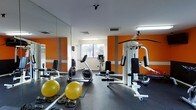 This fitness center is designed to provide a comprehensive workout experience within a compact, purpose-built space. The room is immediately distinguished by its bold orange accent walls contrasted against clean white surfaces, creating an energizing atmosphere conducive to exercise. Full-length mirrors line one wall, visually expanding the space and allowing users to monitor their form during workouts. The flooring features durable rubber matting, offering both safety and comfort underfoot. Equipment includes a multi-station cable and weight machine ideal for full-body strength training, a recumbent exercise bike for low-impact cardio sessions, and an assortment of stability balls and free weights for versatile training options. Natural light enters through a window along the far wall, complementing the overhead fluorescent lighting to keep the space bright and well-illuminated. Ceiling-mounted ventilation ensures comfortable air circulation throughout. The layout is thoughtfully organized to maximize usable floor space while keeping equipment accessible. Overall, the fitness center delivers a practical, well-maintained environment suited for a range of exercise routines, from strength training to cardiovascular and flexibility workouts.
