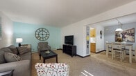 Located at 9112 W Waterford Square S in Greenfield, Wisconsin, this single-floor unit offers a well-organized layout designed for comfortable everyday living. The spacious living room features wall-to-wall carpeting, a soothing teal accent wall, and a open sightline that connects seamlessly to the dining area, creating an inviting, open feel throughout the main living space. Natural light filters in through the windows, brightening the neutral-toned interiors. The galley-style kitchen is efficiently designed with warm honey-toned wood cabinetry lining both sides, white appliances including a range and refrigerator, a stainless steel sink, and tile flooring that contrasts neatly with the carpeted areas. Adjacent to the kitchen, the dining area provides a defined space for meals, illuminated by a pendant light fixture overhead. The bedroom is generously proportioned with plush carpeting, vertical blinds allowing for natural light and privacy, and ample room for substantial furniture arrangements. The full bathroom includes a vanity with wood cabinetry and a tub-shower combination. Throughout the unit, the design maintains a cohesive, practical aesthetic with neutral walls and warm wood tones, making it a versatile and move-in-ready space.