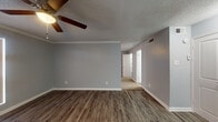This single-story space is designed with a clean, functional layout that connects the main living area, kitchen, and private rooms through an open corridor. The living area is finished with durable wood-style plank flooring in a warm brown tone, complemented by soft gray walls, white crown molding, and a ceiling fan with a built-in light fixture. A textured popcorn ceiling adds a classic touch throughout, and a vertical-blind-covered window allows natural light to filter into the space.

The kitchen is efficiently arranged in a galley-style configuration, featuring white upper and lower cabinetry, dark granite-style countertops, a patterned tile backsplash, and a stainless steel sink. Black appliances, including a range and dishwasher, complete the setup. The kitchen flooring continues the wood-style planks seen in the living area.

A hallway leads to the additional rooms, where carpet flooring begins, providing a softer transition into the private areas of the unit. The bathroom includes a full-size tub and shower combination with white tile surround. Overall, the space presents a cohesive, neutral palette with practical finishes throughout.