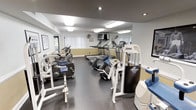 This fitness center is a dedicated single-floor exercise facility offering a comprehensive selection of workout equipment. The space is outfitted with multiple cardio machines, including treadmills, ellipticals, and stationary bikes, alongside strength training equipment from Magnum, providing users with a well-rounded exercise experience.

The room features a neutral color palette with warm yellow-toned lower walls and light gray upper walls, contributing to an inviting yet energizing atmosphere. Recessed and flush-mounted fluorescent lighting ensures the space is well-lit throughout. A large wall mirror along the back enhances the sense of depth and allows users to monitor their form during workouts.

Rubber flooring covers the exercise area, providing both durability and safety underfoot. Natural light filters in through windows fitted with vertical blinds, adding a welcoming brightness to the space. A ceiling fan assists with air circulation, keeping the environment comfortable during intensive sessions. Framed artwork on the walls adds a modest decorative touch. The layout is thoughtfully arranged to allow easy movement between machines while maximizing the use of available floor space.