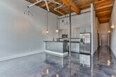 Located at 710 Greenville St in LaGrange, Georgia, this single-floor industrial loft conversion masterfully combines historic mill character with contemporary design. Throughout the space, gleaming polished concrete floors reflect abundant natural light, creating a striking visual continuity from room to room.

The open-concept living area is anchored by oversized multi-pane factory windows set within exposed brick surrounds, flooding the interior with natural light and offering views of the surrounding brick architecture. Soaring ceilings with warm wood tongue-and-groove planking and exposed structural elements reinforce the building's industrial heritage.

The kitchen is a standout feature, outfitted with crisp white shaker cabinetry extending to the ceiling, dark stone countertops, a subway tile backsplash, and a full suite of stainless steel appliances including a side-by-side refrigerator with ice maker, stainless range, and microwave. A peninsula with pendant globe lighting provides additional workspace and casual seating.

The bedroom area offers generous proportions with soaring ceilings and direct access to a walk-in closet. A sliding barn-style door adds an industrial design touch along the corridor. The bathroom features a grey shaker vanity with a dark stone countertop, round mirror, and refined fixtures. The overall ambiance is a seamless fusion of raw, historic industrial elements with clean, modern upgrades.
