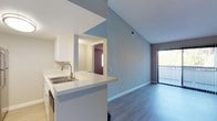 This single-floor residence showcases a thoughtfully designed open-concept layout that seamlessly connects the kitchen and living areas. The kitchen features a peninsula counter with a double stainless steel sink, crisp white cabinetry, and a pass-through window that enhances the open flow between rooms. The light grey countertops and clean finishes give the kitchen a modern, polished aesthetic.

The living area benefits from vaulted ceilings that lend a sense of spaciousness and airiness to the space. Cool-toned grey walls complement the smooth hardwood-style plank flooring that runs throughout, creating a cohesive and contemporary look. A sliding glass door fitted with vertical blinds fills the living area with natural light and provides access to a private balcony, inviting the outdoors in.

A hallway visible from the main living space connects to additional rooms, one of which features a warm, deep accent wall that adds a touch of character and contrast to the otherwise neutral palette. The textured ceiling and clean white baseboards throughout contribute to the unit's well-maintained and move-in-ready condition. Overall, the space offers a bright, functional, and modern living environment with a welcoming ambiance.