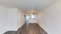 This two-floor space presents a clean, straightforward layout designed for functional everyday living. The main level showcases an open-concept living area that flows directly toward the kitchen, creating a cohesive and connected floor plan. Dark hardwood-style flooring runs throughout the living space, offering a rich contrast against the bright white walls and white baseboards that frame the room. The ceiling is smooth and finished with a ceiling fan fitted with integrated lighting, providing both air circulation and illumination to the space.

Toward the rear of the living area, a kitchen peninsula serves as a natural dividing point between the living and cooking spaces. The kitchen features white cabinetry and receives natural light through a window positioned above what appears to be the sink area, brightening the workspace. A door on the left side of the main level provides access to an additional room or utility space. The staircase leading to the second floor is partially visible near the kitchen, indicating the vertical layout of the home. Overall, the space maintains a neutral, freshly finished aesthetic with a move-in ready appearance.