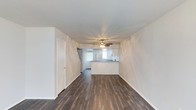 This two-floor space presents a clean, straightforward layout designed for functional everyday living. The main level showcases an open-concept living area that flows directly toward the kitchen, creating a cohesive and connected floor plan. Dark hardwood-style flooring runs throughout the living space, offering a rich contrast against the bright white walls and white baseboards that frame the room. The ceiling is smooth and finished with a ceiling fan fitted with integrated lighting, providing both air circulation and illumination to the space.

Toward the rear of the living area, a kitchen peninsula serves as a natural dividing point between the living and cooking spaces. The kitchen features white cabinetry and receives natural light through a window positioned above what appears to be the sink area, brightening the workspace. A door on the left side of the main level provides access to an additional room or utility space. The staircase leading to the second floor is partially visible near the kitchen, indicating the vertical layout of the home. Overall, the space maintains a neutral, freshly finished aesthetic with a move-in ready appearance.