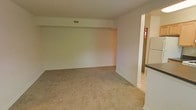 This single-floor residence offers a straightforward and functional layout designed for comfortable everyday living. The main living area is spacious and open, featuring wall-to-wall neutral-toned carpeting that lends a warm, understated feel to the room. Smooth, light-colored walls and a clean ceiling with a central HVAC vent contribute to the space's simple, uncluttered aesthetic. A subtle architectural beam detail runs along the upper portion of the main wall, adding a touch of structural character to the otherwise minimalist design. The living area flows seamlessly into the kitchen, which is accessible through an open entryway. The kitchen features tile flooring, light-toned cabinetry, a dark countertop, and a full-size refrigerator, providing a practical and efficient cooking space. The transition between the carpeted living area and the tiled kitchen is clearly defined, offering a natural separation between the two functional zones. Overall, the space presents a clean, neutral palette throughout, making it highly adaptable for a variety of furnishing styles and personal preferences. The layout prioritizes open space and ease of movement, making it a practical and livable environment.