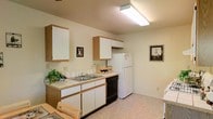 This single-floor residential space is designed with practicality and comfort in mind. The kitchen serves as a central feature, showcasing white lower and upper cabinetry accented with warm wood-tone trim, offering ample storage for everyday needs. Tile countertops line the workspace, complemented by a stainless steel double-basin sink positioned beneath the upper cabinets. The kitchen is equipped with essential appliances, including a black dishwasher and a full-size white refrigerator, making the space move-in ready. A range hood is integrated above the cooking area, contributing to the kitchen's functional layout. Overhead, a flush-mounted fluorescent light fixture provides bright, even illumination throughout the space. The light-toned vinyl flooring adds a clean, cohesive look that ties the room together. Neutral, warm-white walls keep the atmosphere open and inviting, while framed wall art adds a subtle decorative touch. A small wine rack and potted greenery bring personality to the kitchen, enhancing its welcoming ambiance. The overall design reflects a straightforward, well-organized single-floor layout suited for comfortable everyday living.