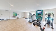 Located at 6317 Robinhood Lane NW in Huntsville, Alabama, this fitness center offers a generously sized, single-floor workout space designed to support a comprehensive exercise routine. The interior features light wood-style flooring throughout, white walls accented by a bold teal feature wall, and recessed lighting that creates a clean, bright atmosphere.

The space is equipped with multiple treadmills, a stationary spin bike, and a dual-cable functional trainer, providing a well-rounded selection of cardio and strength training options. A dumbbell rack with accompanying kettlebells and an adjustable workout bench support free weight training, while full-length mirrors aid in form and technique. A wall-mounted flat-screen television adds an entertainment element to the cardio area.

Natural light enters through windows and a glass door that opens directly to an outdoor pool area, offering views of the surrounding community. A dual water fountain station is mounted along one wall, accompanied by a sanitation station for equipment hygiene. The open floor plan allows for flexible use of the space, including stretching or bodyweight exercises. The fitness center is finished with safety features including a fire extinguisher and clearly marked emergency exits.