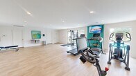 Located at 6317 Robinhood Lane NW in Huntsville, Alabama, this fitness center offers a generously sized, single-floor workout space designed to support a comprehensive exercise routine. The interior features light wood-style flooring throughout, white walls accented by a bold teal feature wall, and recessed lighting that creates a clean, bright atmosphere.

The space is equipped with multiple treadmills, a stationary spin bike, and a dual-cable functional trainer, providing a well-rounded selection of cardio and strength training options. A dumbbell rack with accompanying kettlebells and an adjustable workout bench support free weight training, while full-length mirrors aid in form and technique. A wall-mounted flat-screen television adds an entertainment element to the cardio area.

Natural light enters through windows and a glass door that opens directly to an outdoor pool area, offering views of the surrounding community. A dual water fountain station is mounted along one wall, accompanied by a sanitation station for equipment hygiene. The open floor plan allows for flexible use of the space, including stretching or bodyweight exercises. The fitness center is finished with safety features including a fire extinguisher and clearly marked emergency exits.